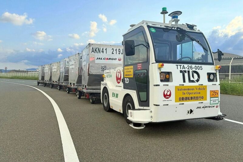 Autonomous cargo vehicle pulling a line of silver shipping containers on a curved highway The truck has operation stickers and beacon lights on top