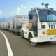 Autonomous cargo vehicle pulling a line of silver shipping containers on a curved highway The truck has operation stickers and beacon lights on top