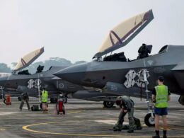 Row of gray military fighter jets with open canopies parked on a tarmac while ground crew in safety vests perform preflight tasks