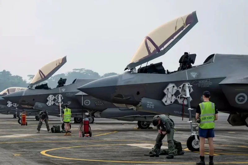 Row of gray military fighter jets with open canopies parked on a tarmac while ground crew in safety vests perform preflight tasks