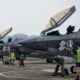 Row of gray military fighter jets with open canopies parked on a tarmac while ground crew in safety vests perform preflight tasks
