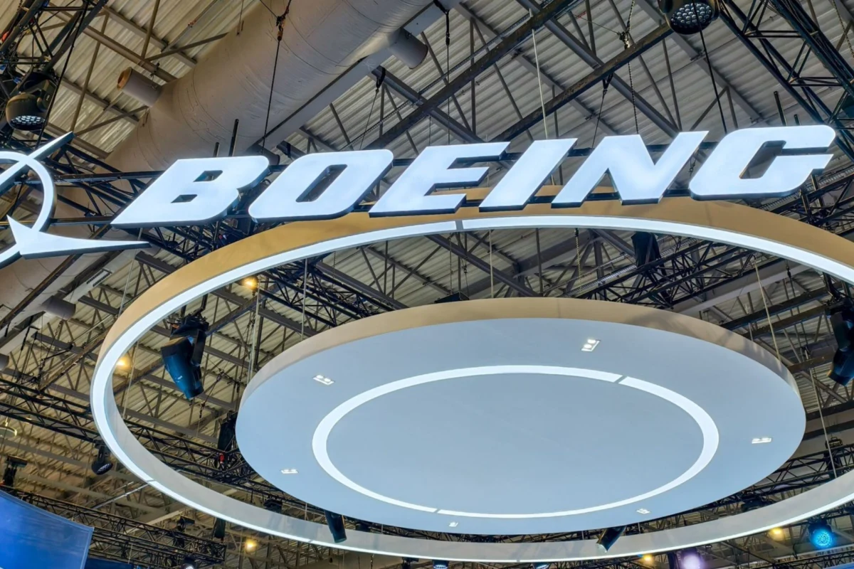 Boeing Q3 2026 record backlog 737 7 and 737 10 certified Bright illuminated BOEING logo suspended from a metal exhibition hall ceiling with circular white rings beneath the sign at a trade show