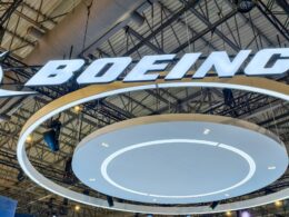 Bright illuminated BOEING logo suspended from a metal exhibition hall ceiling with circular white rings beneath the sign at a trade show