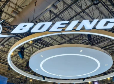 Bright illuminated 'BOEING' logo suspended from a metal exhibition hall ceiling with circular white rings beneath the sign, at a trade show.