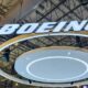 Bright illuminated BOEING logo suspended from a metal exhibition hall ceiling with circular white rings beneath the sign at a trade show