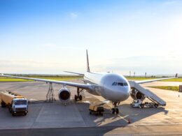 White commercial airplane parked at a gate connected to a jet bridge with ground support vehicles nearby on the tarmac