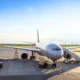 White commercial airplane parked at a gate connected to a jet bridge with ground support vehicles nearby on the tarmac