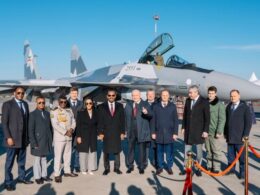 Group of officials and military personnel posing in front of a gray fighter jet with an open cockpit on a sunny airfield