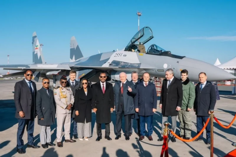 Group of officials and military personnel posing in front of a gray fighter jet with an open cockpit on a sunny airfield