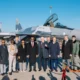 Group of officials and military personnel posing in front of a gray fighter jet with an open cockpit on a sunny airfield