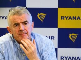 Older man with glasses perched on his head thoughtful expression hands near chin seated in front of a blueyellow Ryanair backdrop