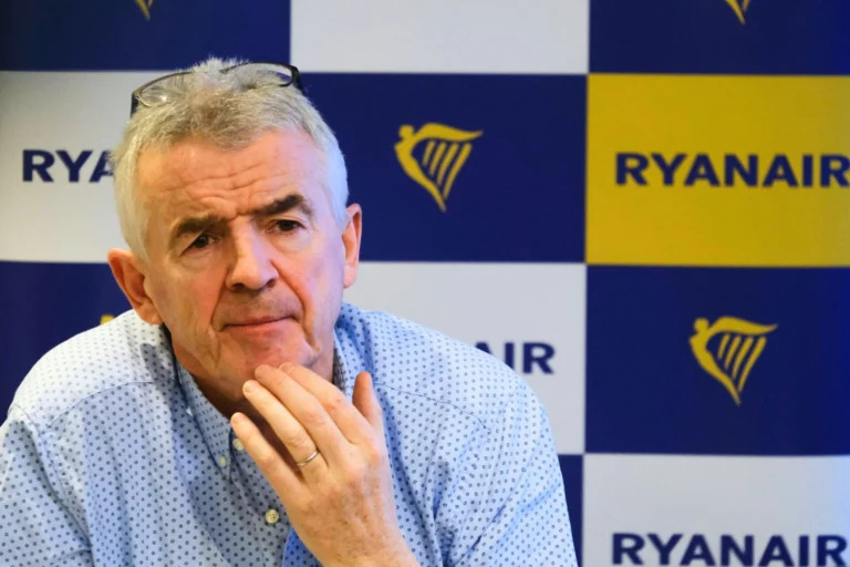 Older man with glasses perched on his head thoughtful expression hands near chin seated in front of a blueyellow Ryanair backdrop