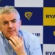 Older man with glasses perched on his head thoughtful expression hands near chin seated in front of a blueyellow Ryanair backdrop