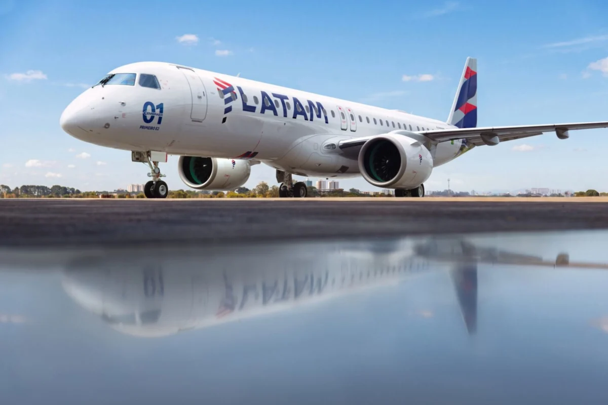 LATAM Brazil takes first E195 E2 to broaden domestic network Side view of a white LATAM commercial jet on a runway under a blue sky engines visible and reflection on the wet tarmac nearby