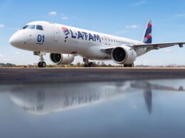 Side view of a white LATAM commercial jet on a runway under a blue sky engines visible and reflection on the wet tarmac nearby