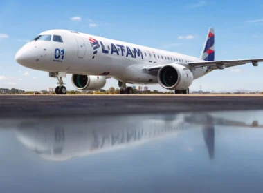 Side view of a white LATAM commercial jet on a runway under a blue sky, engines visible and reflection on the wet tarmac nearby.