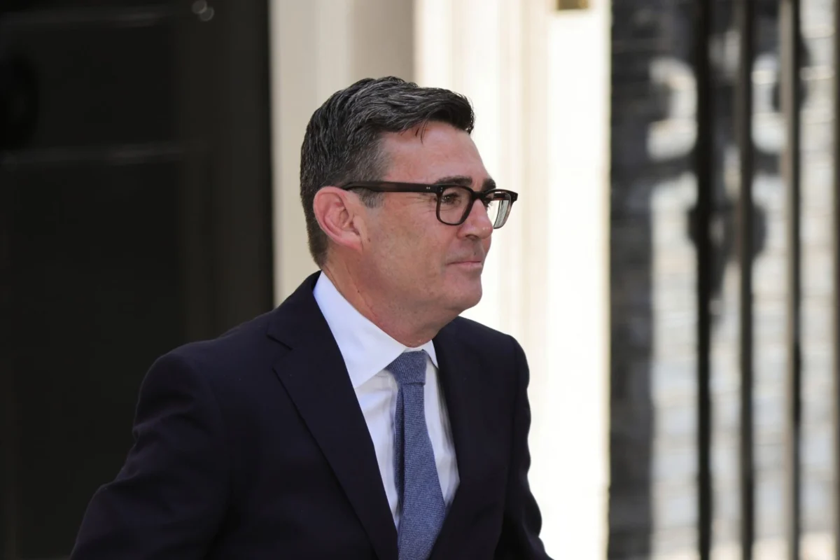 Man in a dark suit and blue tie wearing glasses standing outside near a light colored wall