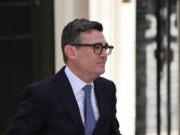 Man in a dark suit and blue tie wearing glasses standing outside near a light colored wall