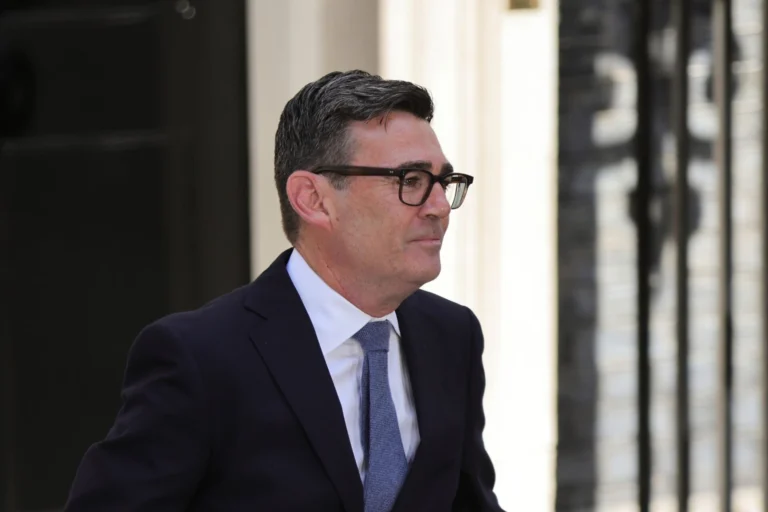 Man in a dark suit and blue tie wearing glasses standing outside near a light colored wall