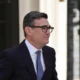 Man in a dark suit and blue tie wearing glasses standing outside near a light colored wall