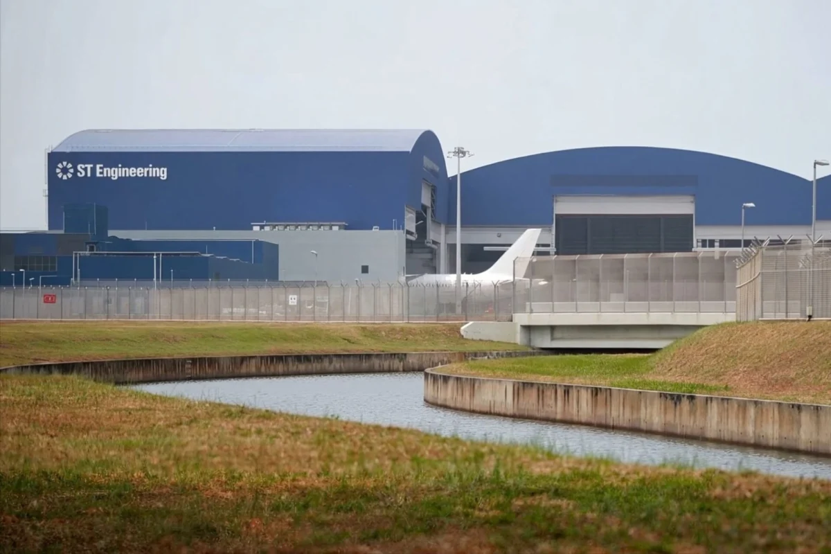 ST Engineering opens AI driven airframe MRO hub in Singapore Blue industrial building labeled ST Engineering behind a perimeter fence with a canal in the foreground