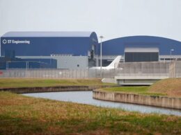 Blue industrial building labeled ST Engineering behind a perimeter fence with a canal in the foreground