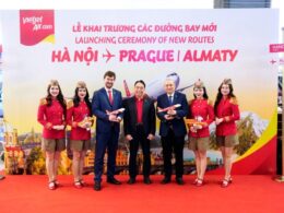 Six people stand on a red carpet in front of a banner for VietJet Airs new routes HanoiPragueAlmaty four flight attendants in red uniforms and hats flank two men in suits each holding a model airplane