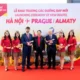 Six people stand on a red carpet in front of a banner for VietJet Airs new routes HanoiPragueAlmaty four flight attendants in red uniforms and hats flank two men in suits each holding a model airplane