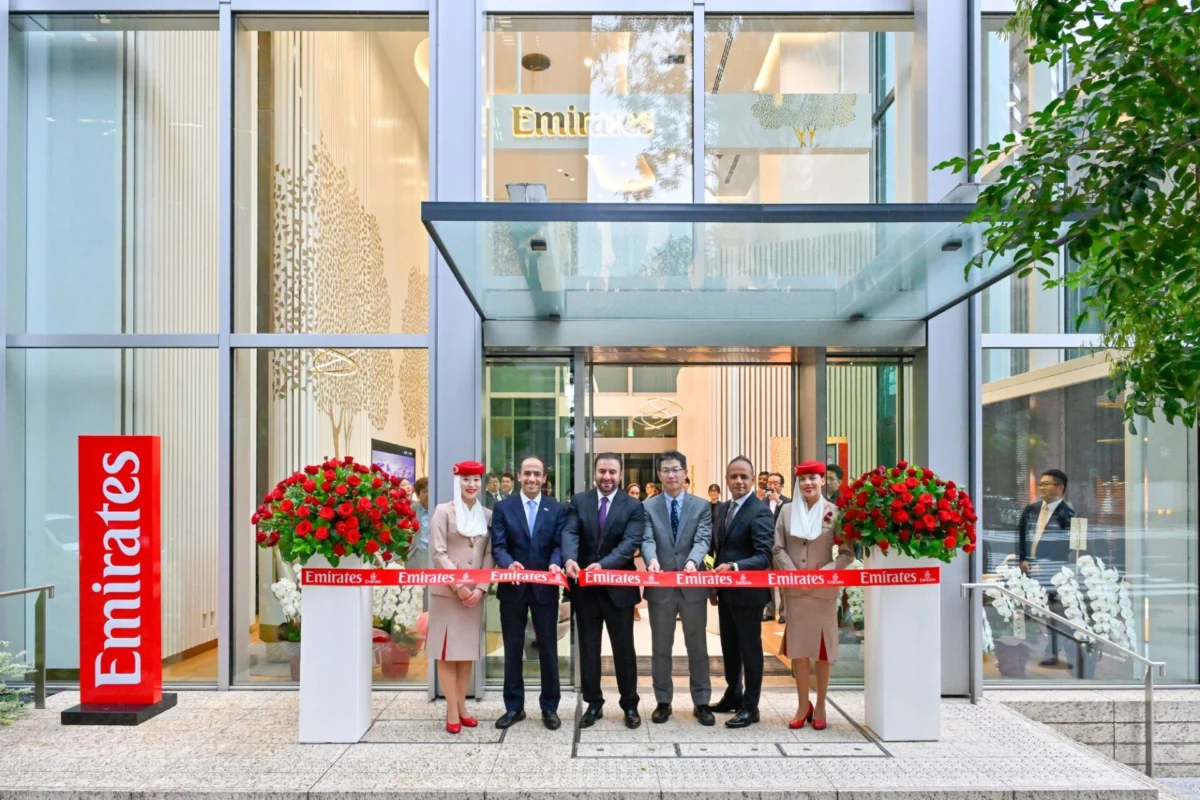 Ribbon cutting ceremony outside a glass Emirates building with staff in business attire and large red flower arrangements outside the entrance