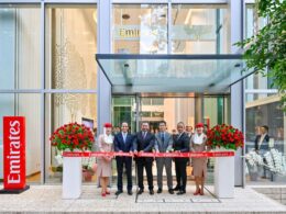 Ribbon cutting ceremony outside a glass Emirates building with staff in business attire and large red flower arrangements outside the entrance