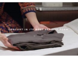 Folded gray garments on a bed with a person folding in the background text overlay reads Comfort in its most refined form