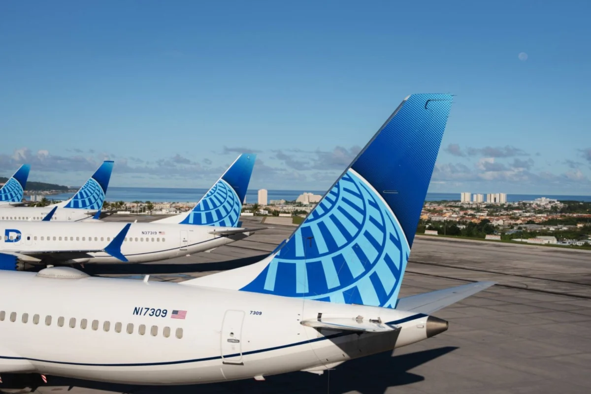 Several large commercial airplanes with blue patterned tails parked on a sunlit airport tarmac coastline visible in the distance