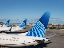 Several large commercial airplanes with blue patterned tails parked on a sunlit airport tarmac coastline visible in the distance