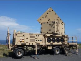 Beige mobile military radarcommunications unit on a trailer with a pyramid shaped perforated top outdoors by the coast