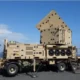 Beige mobile military radarcommunications unit on a trailer with a pyramid shaped perforated top outdoors by the coast