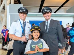 Two airline pilots in uniform pose with a smiling child in front at an indoor event The child holds hands together in a prayer like gesture as crowds mingle in the background