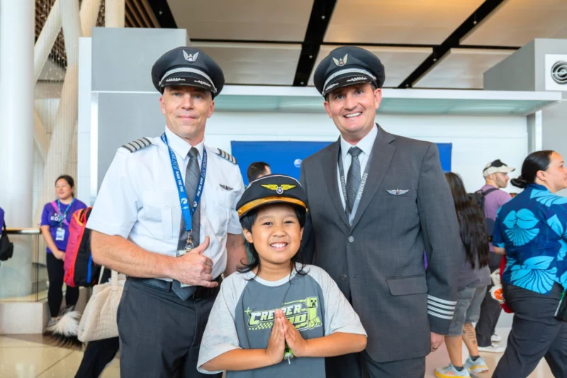 Two airline pilots in uniform pose with a smiling child in front at an indoor event The child holds hands together in a prayer like gesture as crowds mingle in the background