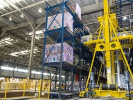 Industrial warehouse showing tall blue pallet racks with wrapped pallets and a large yellow automated lifting crane nearby in operation