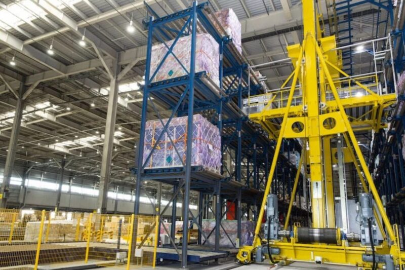 Industrial warehouse showing tall blue pallet racks with wrapped pallets and a large yellow automated lifting crane nearby in operation