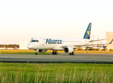 White Alliance airliner taxiing on the airport taxiway with a blue tail and yellow logo, other planes in the background.