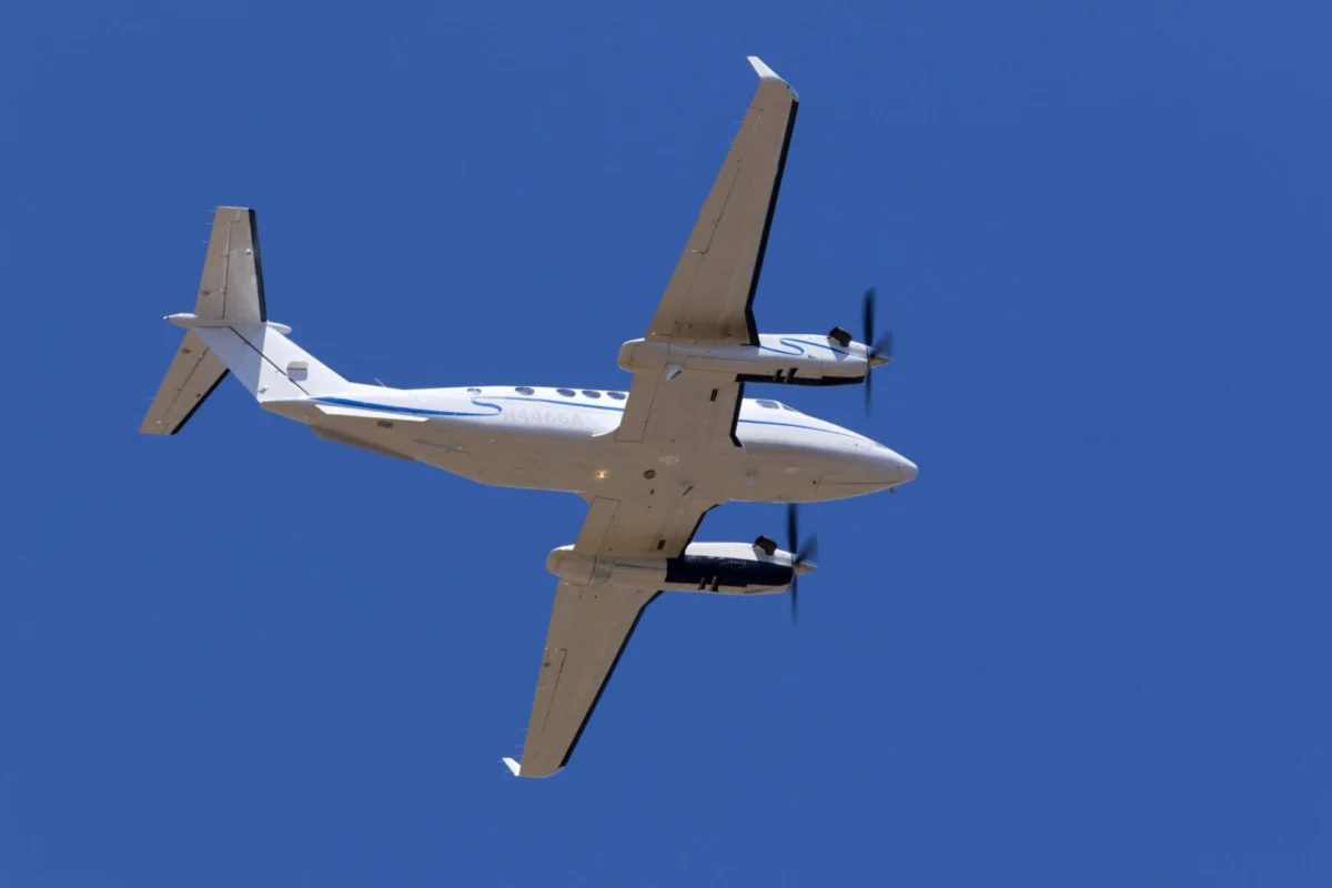 Bottom up view of a white twin engine propeller airplane flying against a clear blue sky