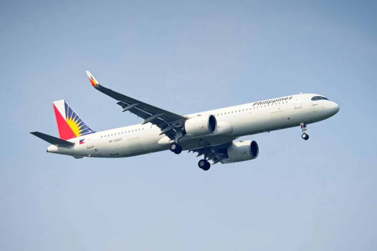 White passenger jet labeled Philippines climbs into a clear blue sky tail adorned with a colorful sun flag