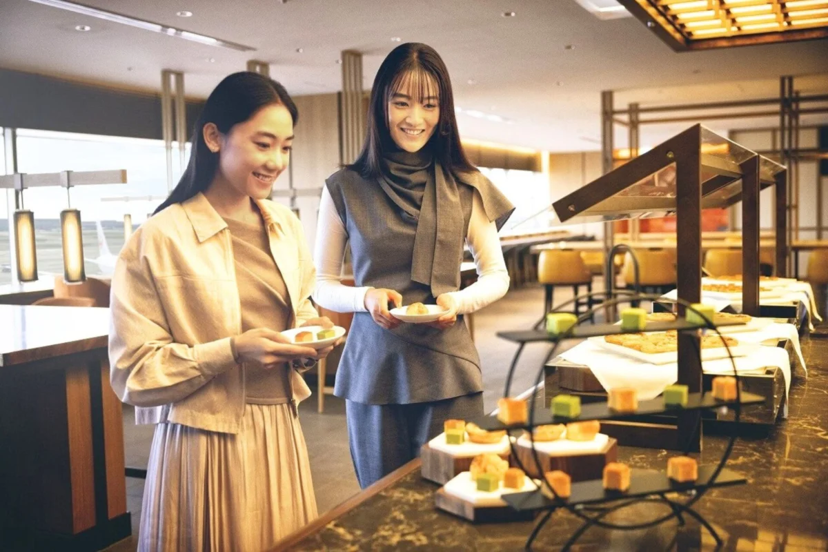 Japan Airlines unveils redesigned domestic lounges at Haneda Two smiling women pick pastries at a self serve buffet plate in hand and a dessert display in front of them