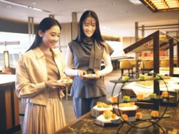 Two smiling women pick pastries at a self serve buffet plate in hand and a dessert display in front of them