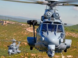 Two gray military helicopters hovering low over a desert like landscape with hills in the background