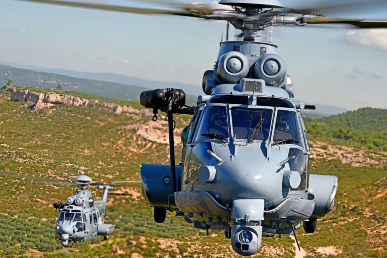 Two gray military helicopters hovering low over a desert like landscape with hills in the background