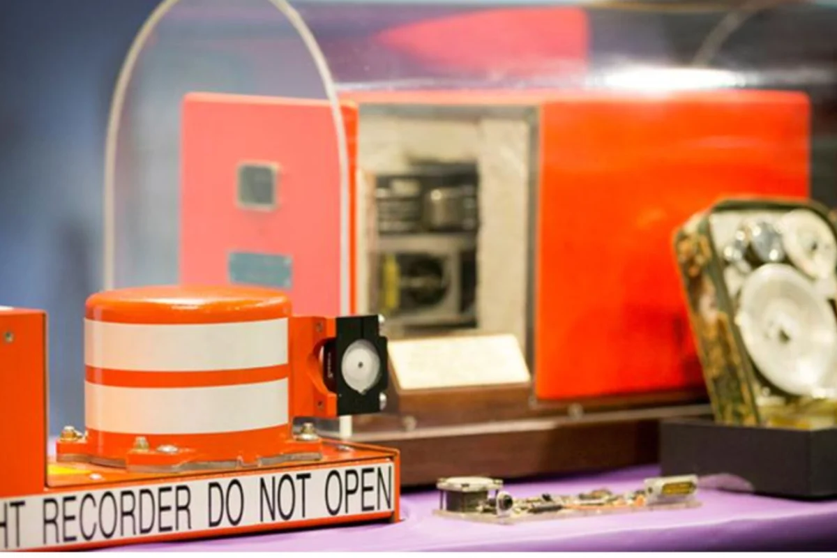 Close up of an orange recording device with a DO NOT OPEN warning on a purple tabletop with machinery in the background