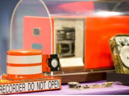Close up of an orange recording device with a DO NOT OPEN warning on a purple tabletop with machinery in the background