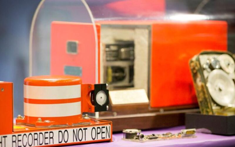 Black box features on coin honoring Australian inventions Close up of an orange recording device with a DO NOT OPEN warning on a purple tabletop with machinery in the background