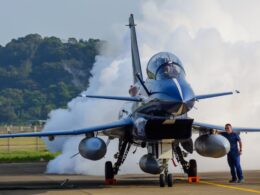 Blue fighter jet on runway with smoke behind it and a ground crew member in blue standing beside the aircraft cockpit canopy visible and orange chocks at the nose gear